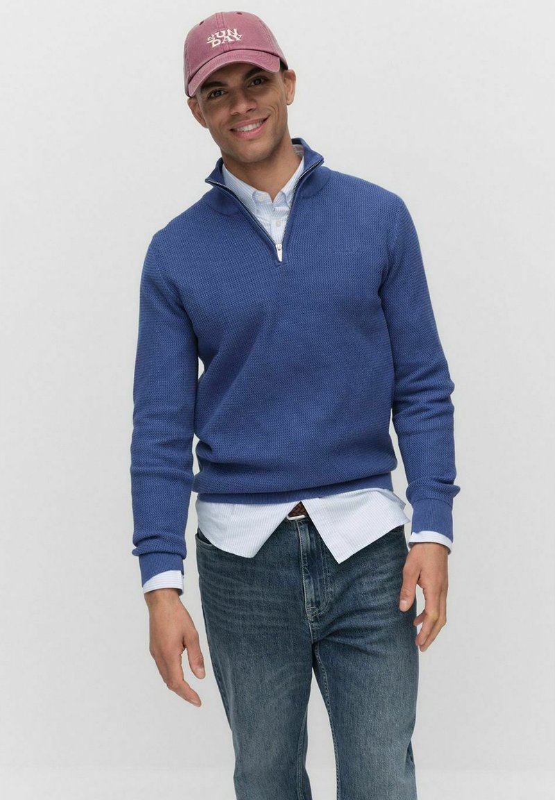 Blå texturerad tröja med halvdäckning och ribbade manschetter, som bärs över en kragblus, matchad med mörka jeans och en rosa mössa.