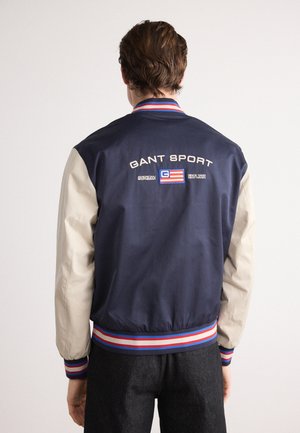Homme portant une veste de sport GANT bleu marine et beige avec des poignets et un ourlet rayés rouge, blanc et bleu, vu de dos.