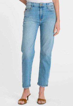 Femme portant un jean taille haute bleu clair avec des revers et des sandales à talons ouvertes marron, debout devant un fond blanc uni.