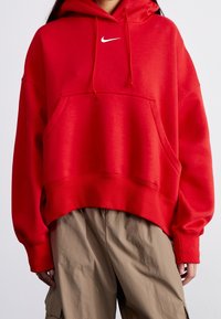 Röd oversized hoodie gjord av bomullsmaterial. Har en framficka av kangurustil och en vit logotyp på bröstet. Texturerade ribbade muddar.