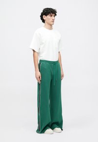 Bărbat stând îmbrăcat cu un tricou alb și pantaloni de trening verzi lejeri cu dungi negre și albe pe lateral, purtând adidași albi, pe un fundal simplu.