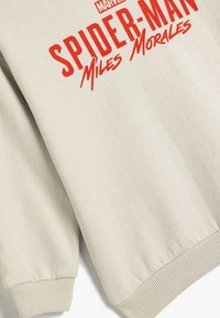Beżowa bluza z czerwonym napisem "SPIDER-MAN Miles Morales" umieszczonym w wyraźnym miejscu. Żebrowane mankiety i detal w dolnej części. Gładka tekstura materiału.
