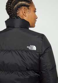 Black puffer jacket with a high collar, featuring a white logo on the upper back, smooth texture, and quilted design for insulation.
