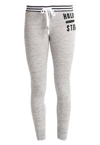 Světlé šedé slim-fit tepláky s černobílým pruhovaným pasem, bílou šňůrkou na stažení a černým textem "HOLLISTER" na horní levé části stehna.