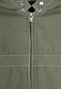 Veste zippée verte avec capuche, fermeture éclair argentée et détails de surpiqûre blanche. Le tissu a une texture lisse et un aspect léger.
