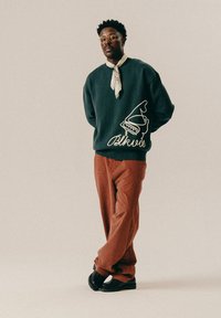 Dark green sweatshirt with white graphic print and text on the side, paired with textured rust-colored pants and black shoes.
