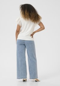 Haut blanc avec des manches en dentelle porté avec un jean bleu clair à jambe large, mettant en valeur une coupe décontractée et un tissu doux. Des talons blancs complètent le look.