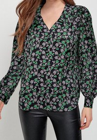 Schwarze Bluse mit grünem und lila Blumenmuster, lange Ärmel, V-Ausschnitt und Knopfverschluss. Kombiniert mit schwarzen, eng anliegenden Hosen.