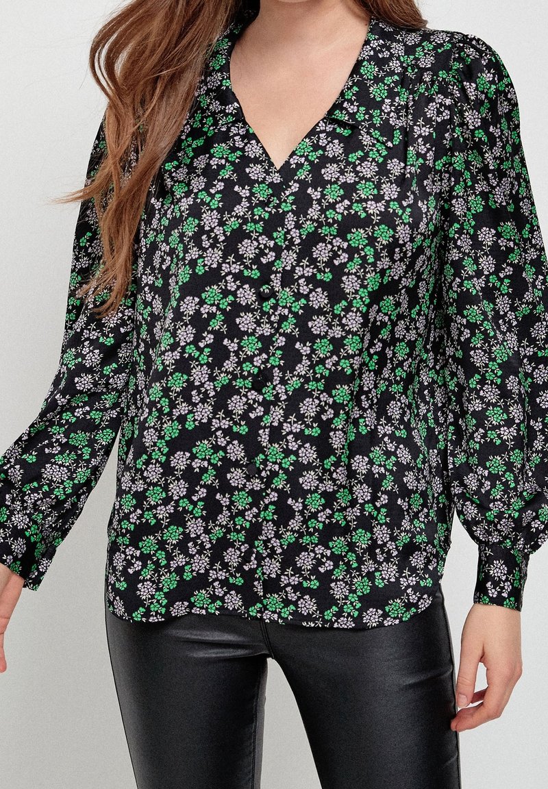 Schwarze Bluse mit grünem und lila Blumenmuster, lange Ärmel, V-Ausschnitt und Knopfverschluss. Kombiniert mit schwarzen, eng anliegenden Hosen.