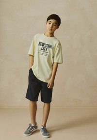 T-shirt oversized jaune clair avec impression "NEW YORK CITY", associé à un short bleu marine foncé et des baskets grises avec des accents blancs.