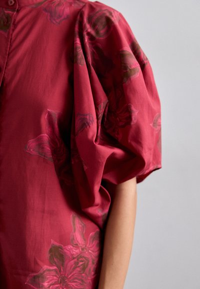 Red blouse with short puff sleeves, featuring floral patterns in darker tones. Made from smooth fabric, it has a buttoned neckline.