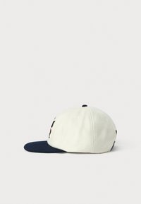 Hvid og marineblå baseballkasket med en flad marineblå skygge, broderet sort og rød design på den synlige side, sat mod en hvid baggrund.
