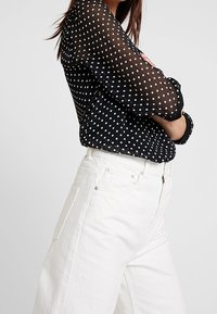 Schwarze, transparente Bluse mit weißen Punkten, langen Ärmeln und abgerundetem Saum, kombiniert mit weißen Jeans mit weitem Bein und Seitentaschen.
