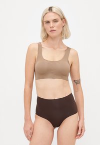 Woman with blonde hair wearing a beige sports bra and high-waisted dark brown underwear, standing against a plain white background.
