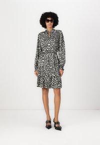 Robe à motifs noir et blanc avec des manches longues, un col à nouer, une taille élastique et un ourlet à volants. Associée à des chaussures à talons noires.
