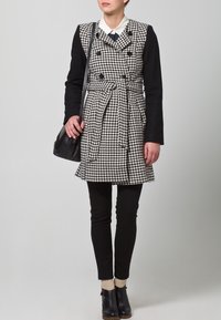 Musta-valkoinen houndstooth-takki, jossa on kaksirivinen muotoilu, vyötäraha, pitkät mustat hiat ja mustat kapeat housut. Täydentää musta laukku.