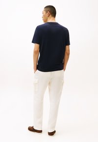 Mann trägt ein marineblaues T-Shirt, weiße Cargo-Hose und braune Schuhe, steht mit den Händen in den Taschen und ist mit dem Rücken zum Betrachter vor einfachem Hintergrund gedreht.