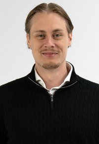 Jonge man met lichtbruin haar en verzorgd gezichtshaar, gekleed in een zwarte ritstrui over een witte kraagshirt, die iets glimlacht tegen een witte achtergrond.