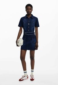 Sukienka koszulowa z denimu w ciemnym kolorze, z krótkimi rękawami, zapięciem na guziki i dwoma przednimi kieszeniami. Stylizowana z paskiem, białą torebką i czerwonymi trampkami z białymi akcentami.
