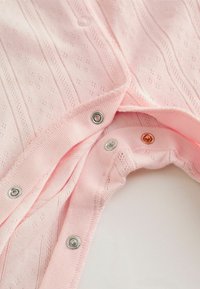 Vêtement tricoté rose clair avec un motif texturé, doté de boutons-pression métalliques en argent et en cuivre, et d'une bordure côtelée douce.