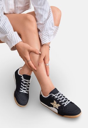 Personne portant des baskets noires avec des lacets blancs et une étoile dorée, assise en tailleur, mains reposant sur le tibia, chemise à manches longues rayée.