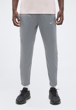 Άνδρας που φοράει γκρι αθλητική φόρμα Nike, λευκές κάλτσες και μαύρα αθλητικά παπούτσια Nike, στέκεται μπροστά σε ένα απλό φωτεινό φόντο.