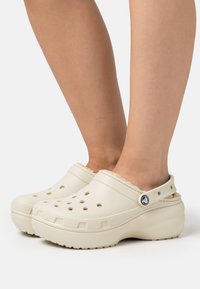 Zuecos beige con un upper texturizado, agujeros de ventilación laterales y un forro suave. Suela de plataforma con un acento de logo circular en la correa.