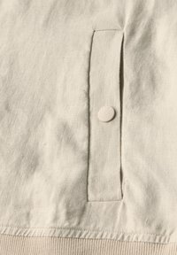 Tessuto beige, probabilmente lino, con una tasca laterale dotata di un flap in tessuto coordinato e un bottone tondo con la scritta "CECIL" in rilievo.