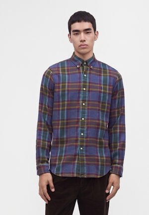 Jeune homme portant une chemise à carreaux bleue, verte et marron à manches longues, debout devant un fond clair uni.