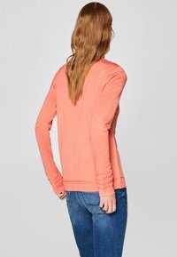 Dlouhý rukáv, korálová blůza vyrobená z měkké látky s volným střihem a žebrovanými manžetami, spárována s tmavě modrými džíny, pohled zezadu.