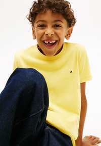 Χαμογελαστό νεαρό αγόρι με σγουρά μαλλιά, φορώντας κίτρινη μπλούζα και σκούρα μπλε τζιν, καθισμένο με το ένα γόνατο σηκωμένο μπροστά σε απλό φόντο.