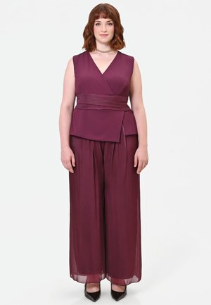 Donna con capelli castani fino alle spalle che indossa un top bordeaux senza maniche avvolgente e pantaloni a gamba larga con scarpe nere a punta su sfondo bianco.
