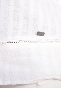Tejido blanco texturizado con rayas horizontales, con un borde de encaje en la parte inferior y una pequeña etiqueta con logotipo plateado.