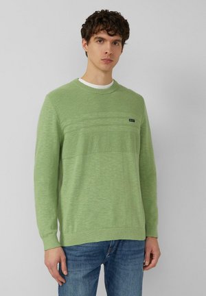 Junger Mann mit lockigem Haar, der einen hellgrünen, strukturierten Strickpullover und blaue Jeans trägt, vor einem schlichten hellgrauen Hintergrund steht.