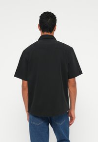 Črna majica s kratkimi rokavi z mehkimi in gladkimi površinami ter klasičnim ovratnikom. Podrobnosti vključujejo hrbtni yoke in ravni rob. Nošena z modrimi kavbojkami.