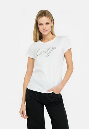 T-shirt en coton blanc avec le texte "Luge" en argent brillant sur le devant. Manches courtes, col rond, associé à un pantalon noir avec des accents cloutés.