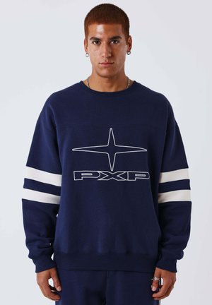 Jonge man met kort rood haar draagt een marineblauwe trui met een wit sterontwerp en de tekst "PXP", staand tegen een eenvoudige lichte achtergrond.