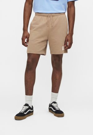 Mies pukeutuneena beigenvärisiin nyörillisiin shortseihin, mustavalkoisiin lenkkareihin, valkoisiin sukkiin ja vaaleansiniseen paitaan yksiväristä taustaa vasten.