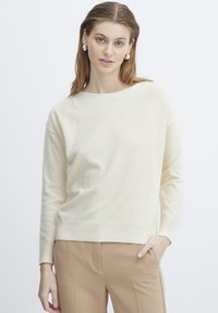 Pull en maille couleur crème avec un col rond, des manches longues, et des bords côtelés. Porté avec un pantalon beige, texture lisse.