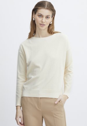 Cremfarbener Strickpullover mit rundem Ausschnitt, langen Ärmeln und ribbed Cuffs und Saum. Getragen mit beigen Hosen, glatte Textur.