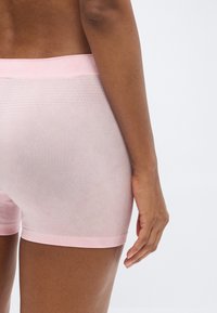 Gros plan sur une personne portant un short sans coutures rose clair, montrant le bas du dos, la hanche et le bras droit sur un fond uni.