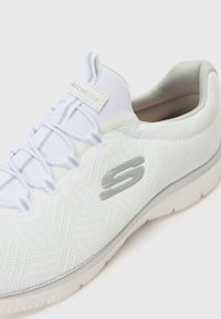 White mesh athletic shoe with a textured surface, elastic laces, and a gray logo on the side. Features a thick, white rubber sole.
