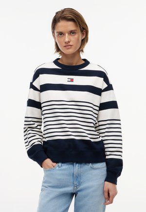 Jeune personne aux cheveux courts portant un sweat-shirt à rayures bleu marine et blanc et un jean bleu clair, debout avec une main dans la poche.