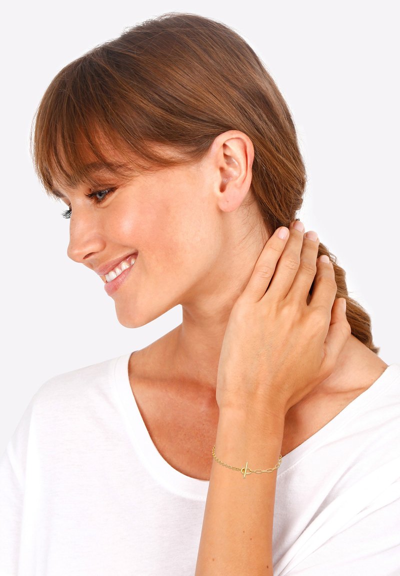 Pulsera de oro con una delicada cadena y un pequeño detalle de cierre central. El modelo lleva una camisa blanca, mostrando la joyería.