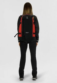Persona con una mochila grande Haglöfs Tight en negro y rojo, de espaldas, vestida con ropa de exterior negra y botas de senderismo.