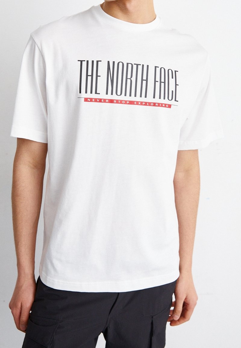 Bílý bavlněný tričko s velkým černým logem "THE NORTH FACE" a červeným akcentem s textem "NEVER STOP EXPLORING" na přední straně. Krátké rukávy.