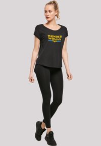 Melnas īsām piedurknēm T-krekls ar "WONDER WOMAN" grafiku dzeltenā un zilā krāsā, apvienots ar melnām legingām un melniem sporta apaviem.