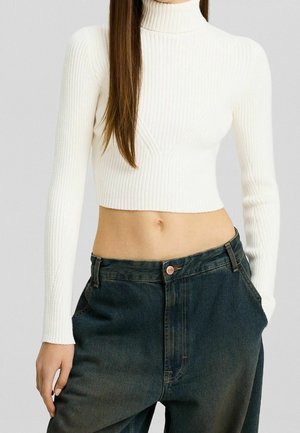 Witte geribbelde turtleneck cropped top met lange mouwen, gecombineerd met donkere denim broek met een getailleerde taille en een voorzak ontwerp.