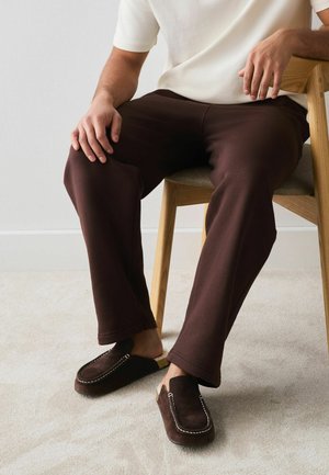 Person sitzt auf einem Holzstuhl und trägt dunkelbraune Hosen, ein weißes Oberteil und dunkelbraune Mokassin-Hausschuhe auf hellem Teppich.