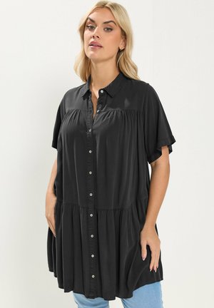 Blondynka w luźnej czarnej sukience na guziki z krótkim rękawem, nosząca jasnoniebieskie dżinsy na jednolitym jasnym tle.